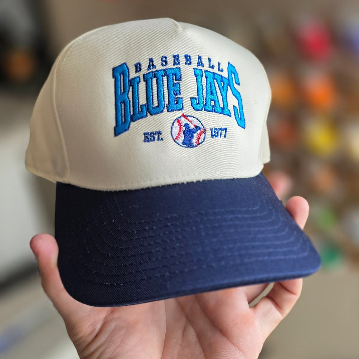 Baseball cap with 'Blue Jays' branding held in front of a colorful background